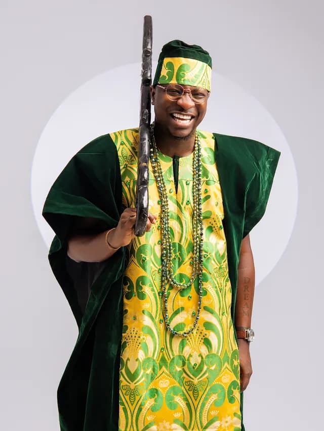 Theo laughing in a green velvet agbada over green and gold damask, holding a carved staff