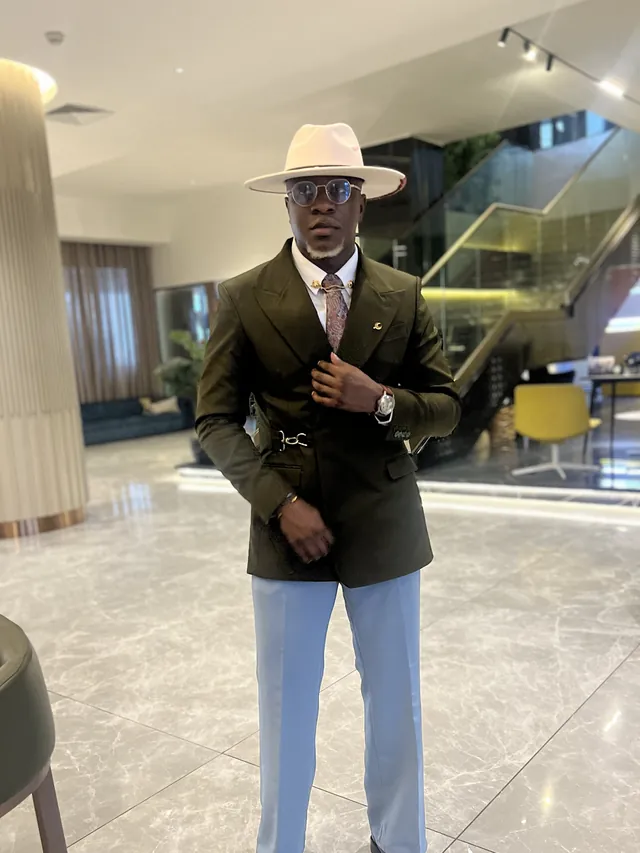 Theo in an olive belted blazer, light blue trousers and cream fedora