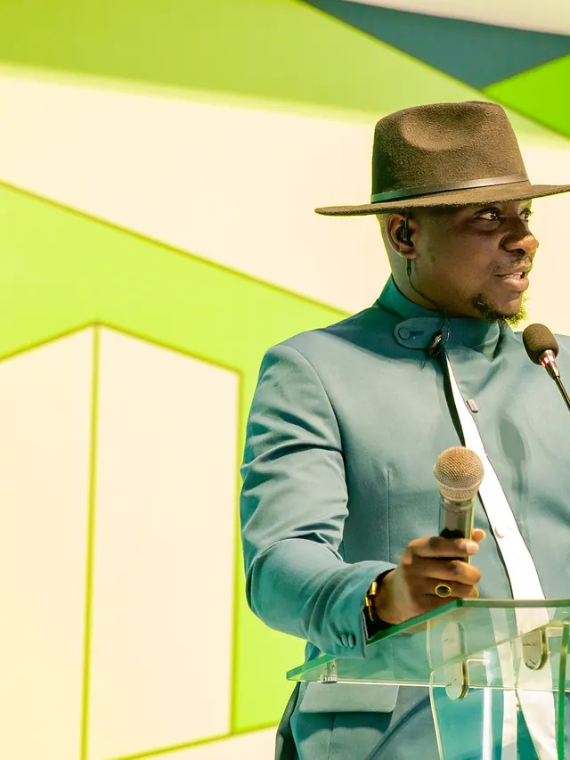 Theo hosting at a glass podium in a teal mandarin-collar suit and brown fedora